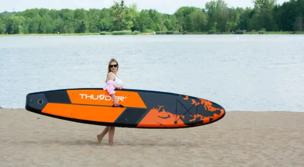 Kobieta z pomarańczowo-czarnymi deskami do pływania z wiosłem na piaszczystej plaży, gotowa do wodnych przygód.