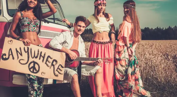 Neo-Hippies mit Gitarre und Schild "Anywhere" vor einem Van in einem Feld.