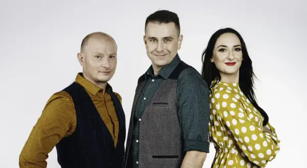 Trzy osoby: łysy mężczyzna w kamizelce, mężczyzna w zielonej koszuli i kamizelce, kobieta w żółtej sukience w grochy.