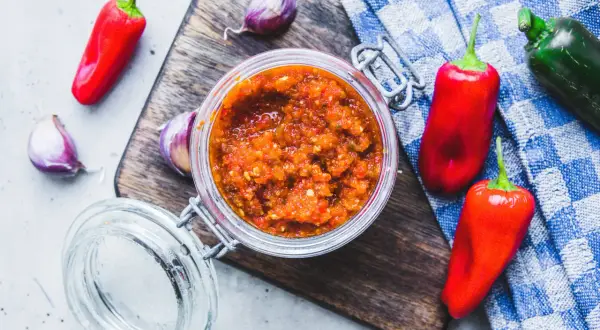 Domowy sos sriracha z papryczek chilli i czosnku w słoiku, otoczony świeżymi papryczkami i czosnkiem na desce.