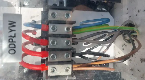 Widok z bliska na zaciski elektryczne z podłączonymi przewodami w różnych kolorach (niebieski, zielono-żółty, brązowy, czarny).