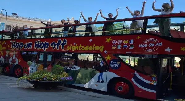 Glada resenärer vinkar från övervåningen på en röd dubbeldäckare med texten "Hop on Hop off sightseeing".