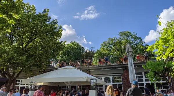 Menschen genießen das Wetter im Golgatha Biergarten. Sonnenschein, grüne Bäume und ein blauer Himmel bilden die Kulisse.