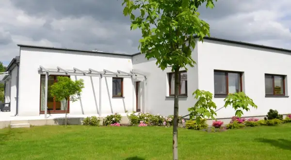 Nowoczesny bungalow z białymi ścianami i dużymi oknami, otoczony zielonym trawnikiem i kwitnącymi krzewami.
