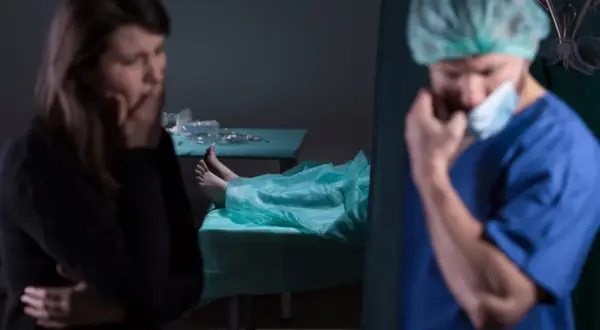 Smutna kobieta i lekarz w masce, obok zakryte ciało na stole. Kiedy sekcja jest obowiązkowa, emocje biorą górę.