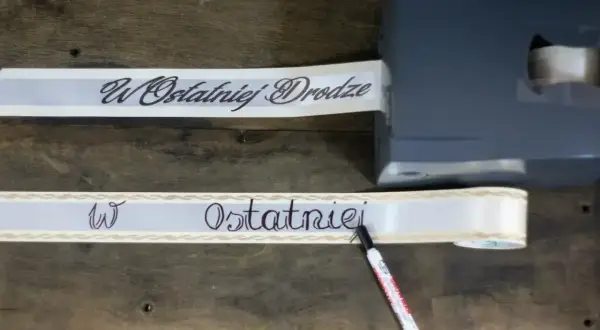 Taśma z napisem "W Ostatniej Drodze" przygotowywana do wieńca. Wzór jest elegancki i symboliczny.