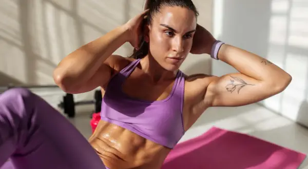 Frau macht Crunches auf einer rosa Matte, um einen flachen Bauch zu bekommen. Sie trägt lila Sportkleidung und hat ein Tattoo auf dem Arm.