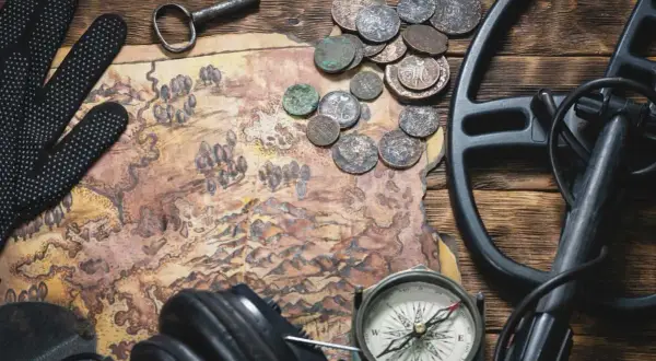 Mapa, monety, klucz, rękawice, kompas i wykrywacz metali na drewnianym stole.