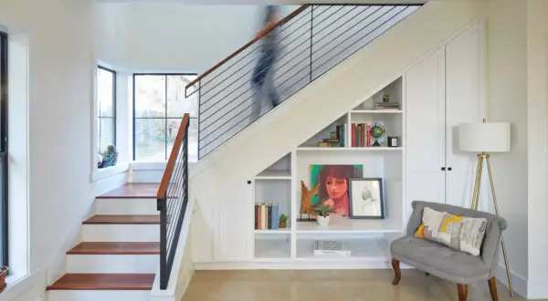 A person ascends a modern staircase, its sleek design contrasting with the built-in bookshelves and storage beneath. This stairwell is a functional art piece.