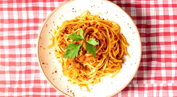 Assiette de spaghetti à la bolognaise, garnie de persil frais, sur une nappe à carreaux rouges et blancs. Une question se pose : peut-on manger des pâtes le soir ?