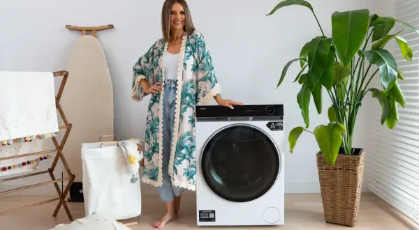 Kobieta w kwiecistej narzutce obok nowoczesnej pralki Haier. Wiele osób szuka pralki Haier opinie, by wybrać najlepszy model.