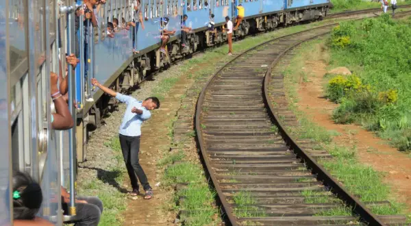 Un tren azul de Sri Lanka avanza por las vías, con pasajeros asomados a las ventanas y otros viajando en los estribos.