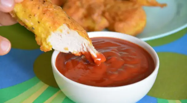 Chrupiący nugget zanurza się w gęstym, czerwonym sosie do nuggetsów. W tle widać talerz z innymi nuggetami.