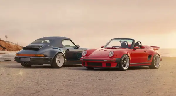 Dos Porsche descapotables, uno rojo y otro azul oscuro, posan en la playa. El **Singer Porsche** rojo destaca con su diseño deportivo y el azul con su capota puesta.