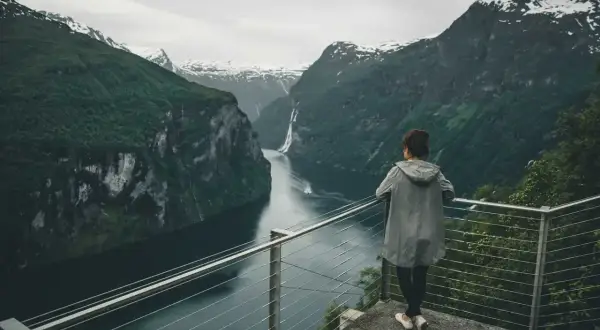 Kobieta podziwia majestatyczne fiordy norweskie. Woda odbija strome, zielone zbocza gór, a w oddali widać wodospad.