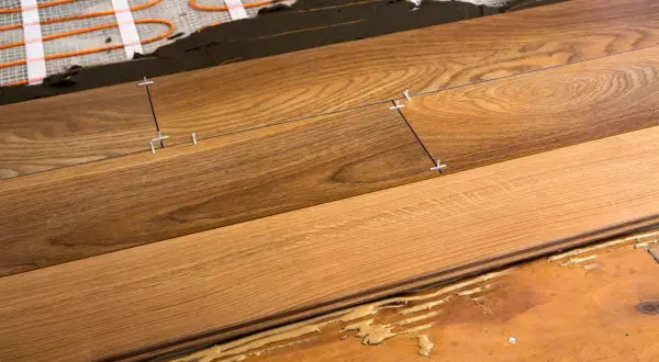 Close-up of plank flooring installation over underfloor heating. Wooden planks are being laid with spacers, showing the adhesive and heating elements beneath.