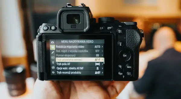 Ręka trzyma bezlusterkowiec pełna klatka, na ekranie menu nagrywania wideo. Ranking aparatów jest w tle.