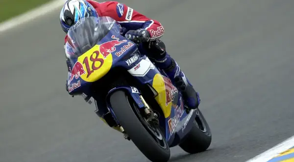 Piloto con casco azul y rojo, traje de carreras Red Bull y moto Yamaha número 18, en plena curva. Jean Michel Bayle en acción.