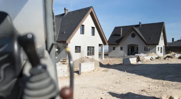 Budowa dwóch domów gospodarczych w słoneczny dzień. Widać fragment maszyny budowlanej na pierwszym planie.