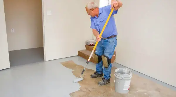 Un homme en bleu peint le sol en béton avec un rouleau. Il porte des genouillères pour peindre le béton confortablement.