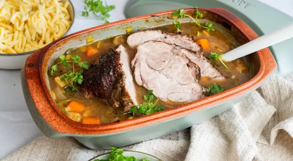 Saftiger schweinebraten im schmortopf, mit Karotten und Lauch, garniert mit Petersilie. Daneben eine Schüssel mit Spätzle.