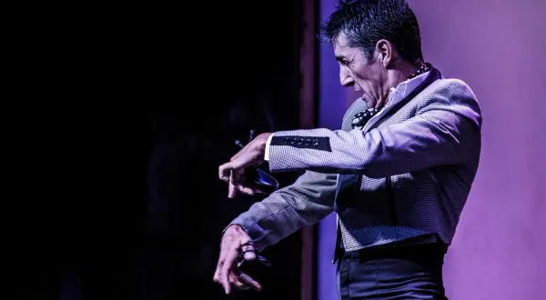 Un hombre con traje a cuadros realiza un apasionado flamenco baile, con las manos extendidas y castañuelas.