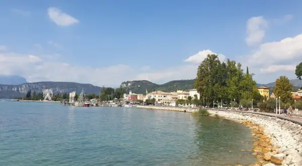 Il lungolago di Bardolino, con il lago azzurro, le barche, le case colorate e le montagne sullo sfondo. Una ruota panoramica si staglia all'orizzonte.