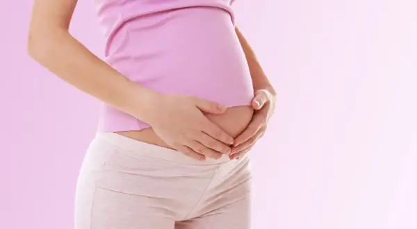 Kobieta w różowej bluzce i beżowych legginsach trzyma dłonie na lekko zaokrąglonym brzuchu. To może być jeden z pierwszych objawów ciąży, jak zmiany w śluzie.