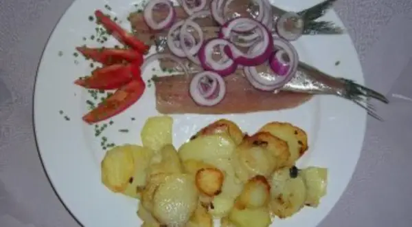 Matjes mit Bratkartoffeln, garniert mit Zwiebelringen und Tomaten. Ein klassisches Gericht, das auf einem weißen Teller serviert wird.