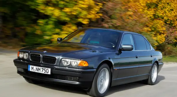 Ciemnoszary BMW E38 Long sunie po drodze, otoczony jesiennymi drzewami w pełnym kolorze.