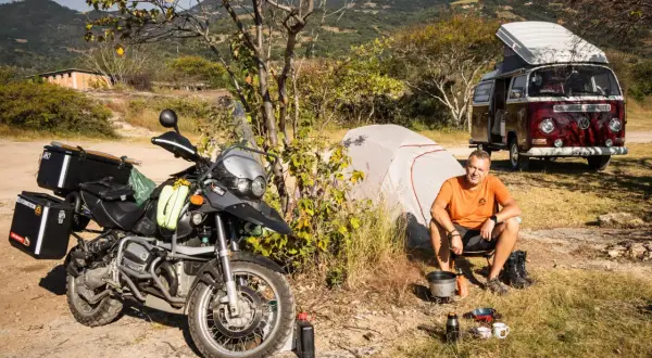 Motocykl, namiot, kamper i mężczyzna siedzący przy ognisku na tle gór.