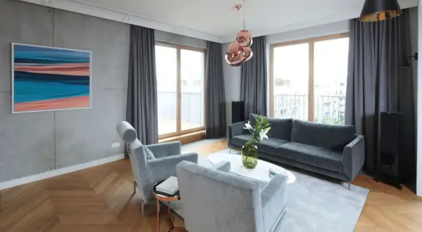Nowoczesny salon z betonową ścianą, drewnianą podłogą w jodełkę, szarymi meblami i miedzianymi lampami. Lamele i beton tworzą industrialny klimat.