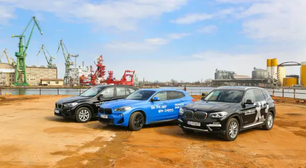 Trzy samochody BMW X: czarny, niebieski i kolejny czarny, stoją na placu budowy z dźwigami w tle.