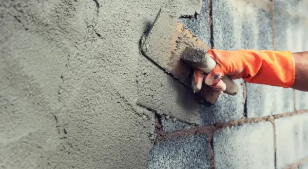 Mano con guanto arancione stende malta da costruzione su un muro di blocchi di cemento con una cazzuola.