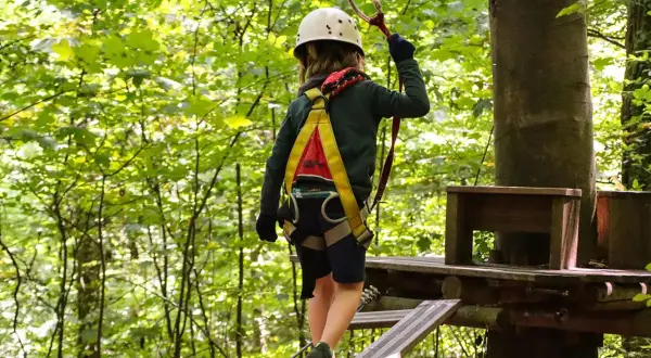 Kind im Hochseilgarten K1 balanciert auf einem schmalen Steg, gesichert durch Klettergurt und Helm.