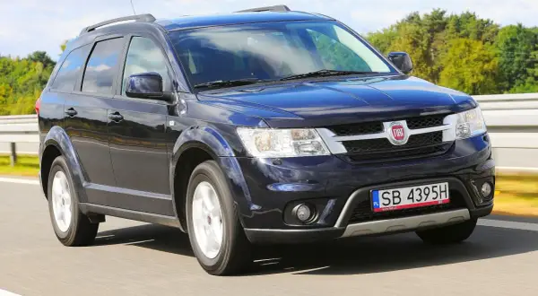 Ciemnoniebieski Fiat Freemont jedzie po drodze, otoczony zielenią i błękitnym niebem.
