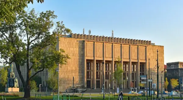 Muzeum Narodowe w Krakowie przy Alei 3 Maja. Monumentalna budowla z kamienia, otoczona zielenią i stojakami z hulajnogami.