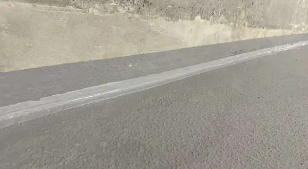 Szara posadzka z widoczną dylatacją wypełnioną białym materiałem, granicząca z betonową ścianą.