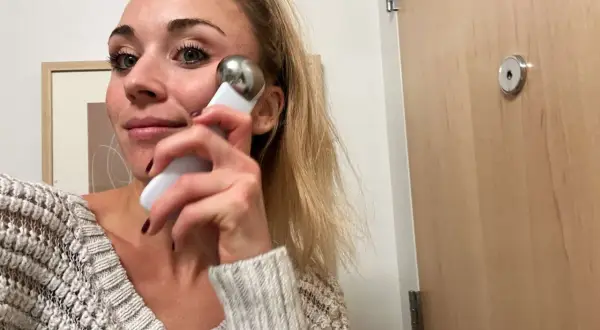 Kvinna med blont hår använder en vit och silverfärgad **ice roller ansikte** för att massera kinden.
