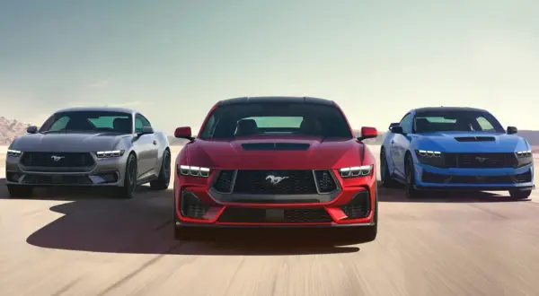 Tre Ford Mustang modeller i rött, grått och blått kör på en ökenväg.
