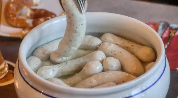 Eine Gabel hebt eine Weißwurst aus einem Topf, um sie zuzubereiten. Daneben liegt eine Brezel.