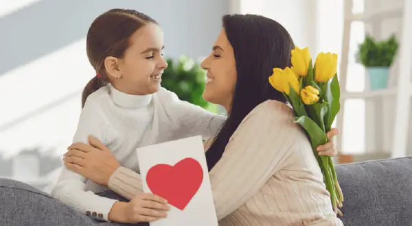 Uśmiechnięta dziewczynka wręcza mamie kartkę z sercem i bukiet żółtych tulipanów. Radosny Dzień Matki UK!