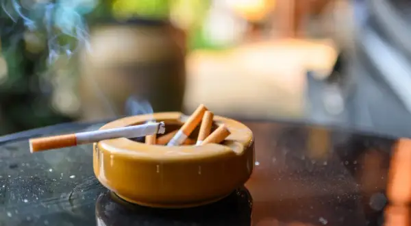 Cenicero lleno de colillas, con un cigarrillo encendido y humo. Un recordatorio de la necesidad de eliminar olor tabaco casa.