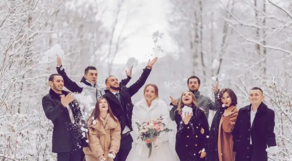 Brautpaar und Gäste werfen Schnee in der Luft. Perfekt für eine Hochzeit im Winter: Was anziehen? Warme Mäntel und festliche Kleidung.