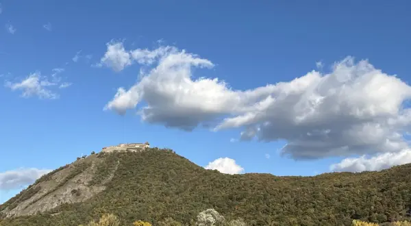 Wyszehrad góruje nad zielonym wzgórzem, z zamkiem na szczycie. Pod nim rozciąga się las, a u podnóża widać rzekę.