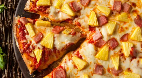 Pyszna hawaiian pizza z kawałkami ananasa i szynki, obficie posypana serem, prezentuje się apetycznie na ciemnym tle.