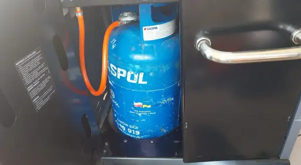 Niebieska butla z gazem marki GASPOL, podłączona wężem do grilla. To odpowiednia butla do grilla gazowego, zapewniająca zasilanie.