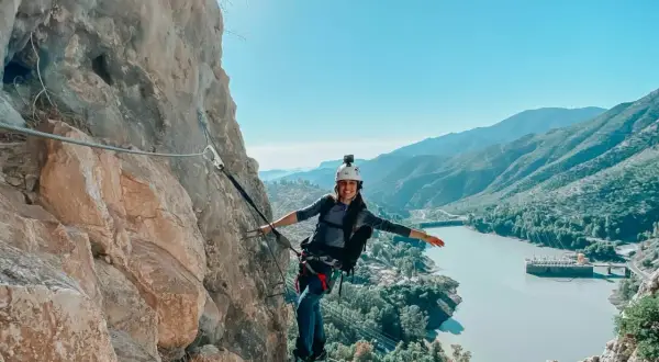 Aventura en el Caminito del Rey. Mujer sonríe mientras recorre la vía ferrata, con un embalse y montañas de fondo.