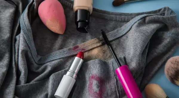 Graues T-Shirt mit Flecken von Lippenstift, Mascara und Foundation. Ein Make-up-Schwamm und Pinsel liegen daneben. Tipps zum Make-up aus Kleidung entfernen.