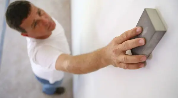 Un homme ponce un mur blanc avec une cale à poncer. C'est une étape essentielle : faut-il poncer un mur avant de le peindre pour un fini parfait ?